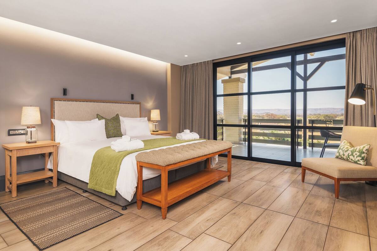 Habitación de hotel con una cama grande, un banco, una silla y vistas al exterior a través de un ventanal.