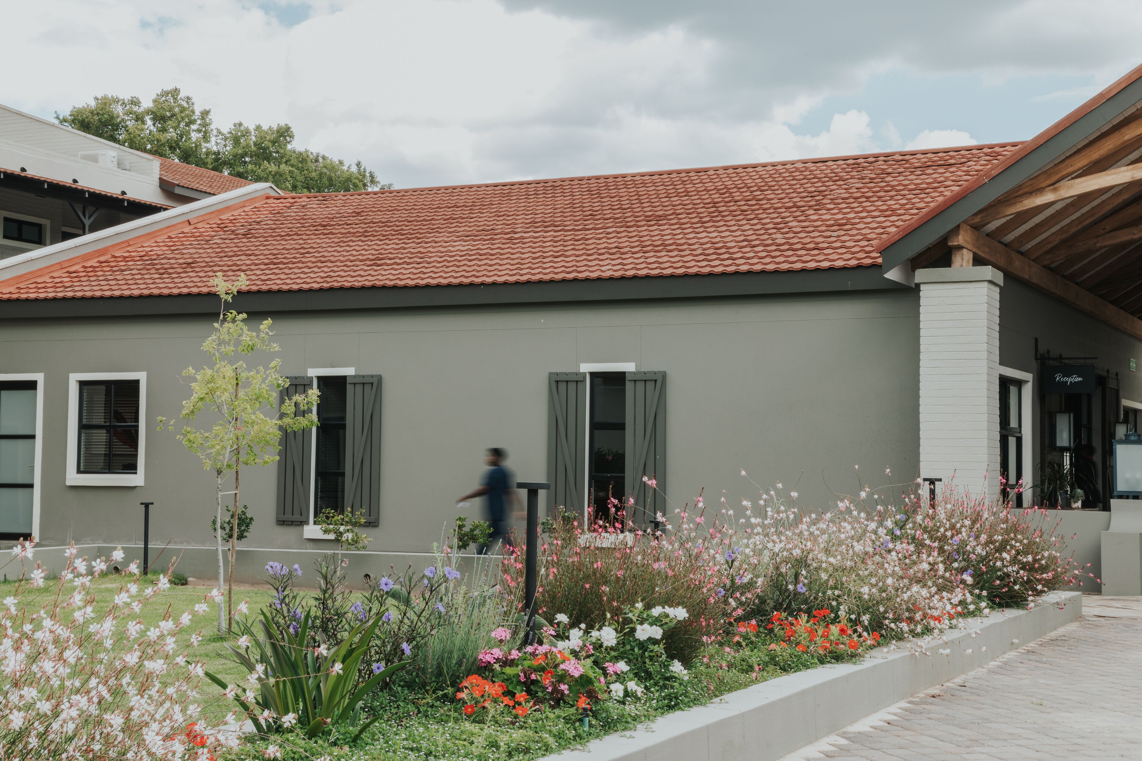 A modern building with a red-tiled roof, grey walls, and a garden with various flowers.