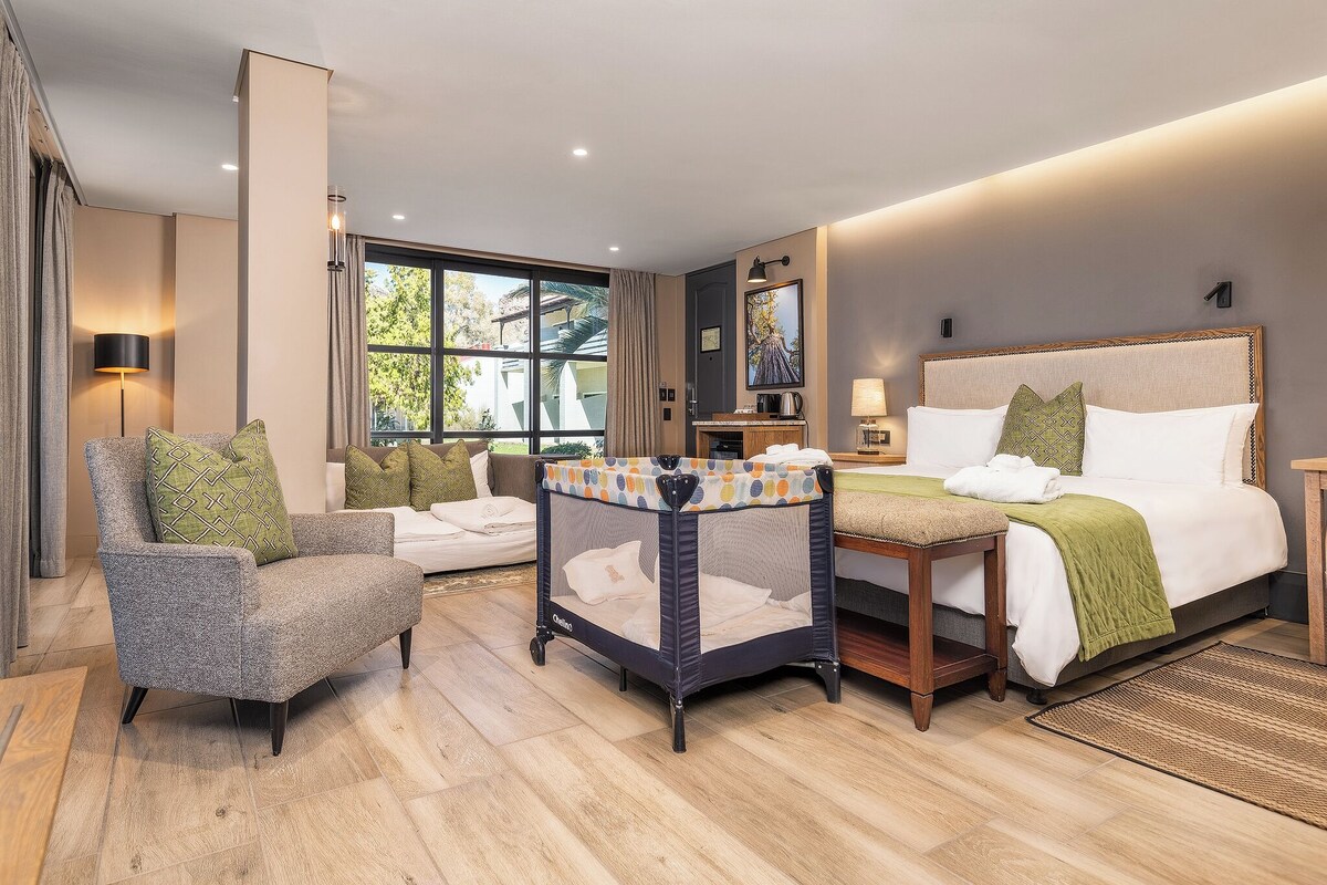 A hotel room with a bed, a chair, a small playpen, a desk, and a window.