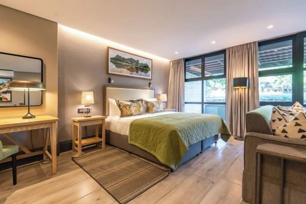 Habitación de hotel con cama, mesitas de noche, escritorio, silla y un amplio ventanal con cortinas.