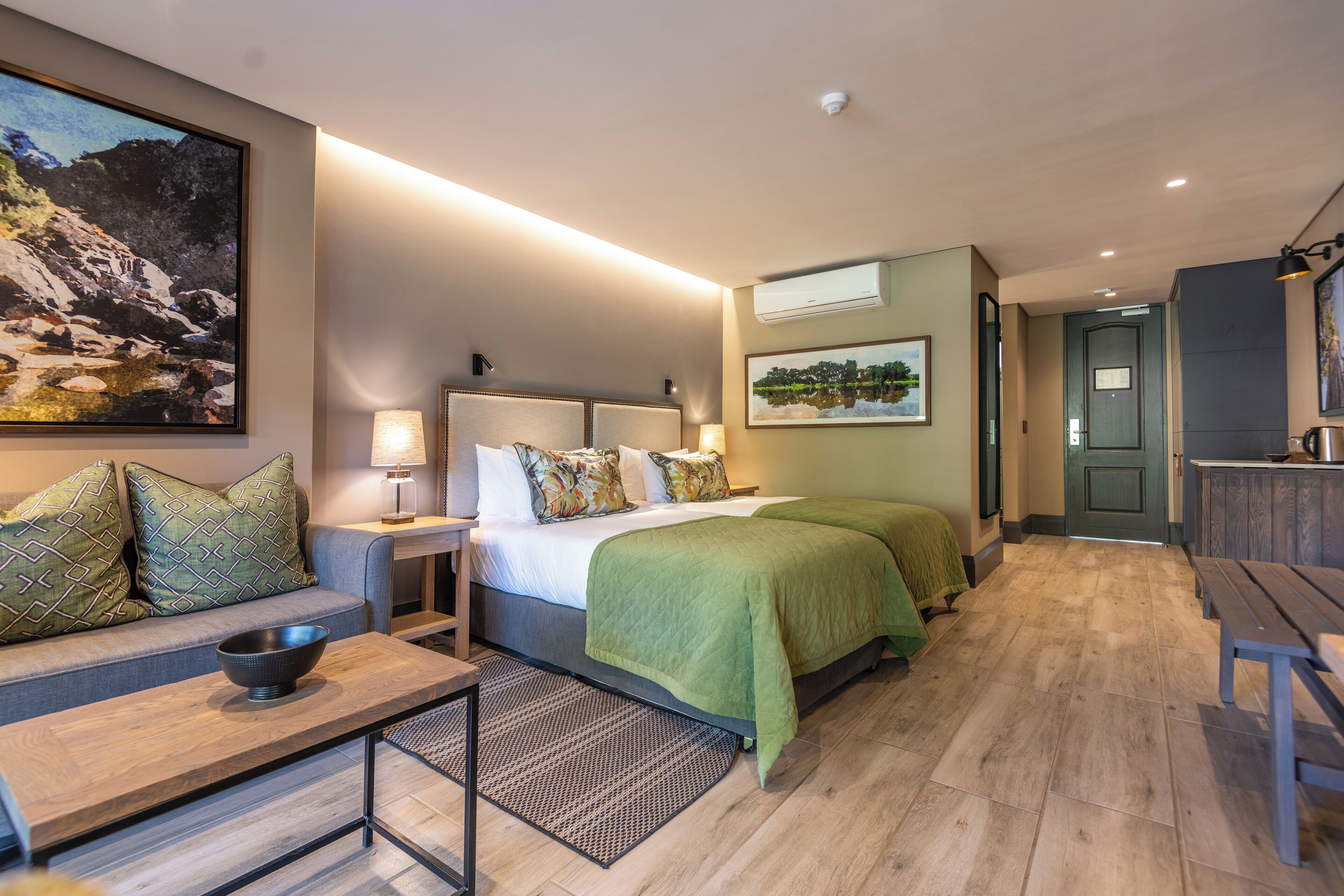 A hotel room with a bed, a sofa, a table with a bowl, a lamp, and a painting on the wall.