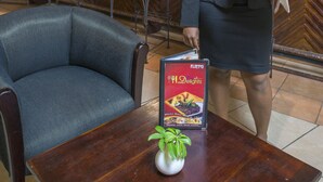 A bar area with wooden furniture, a bar counter with bottles, and a menu stand. - New Ambassador Hotel (Harare)