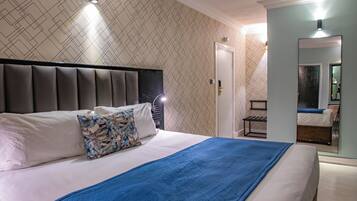 Un lit bien fait, avec une couverture bleue et un coussin à motifs floraux, un mur orné d’un motif géométrique et un miroir qui reflète une partie de la pièce.