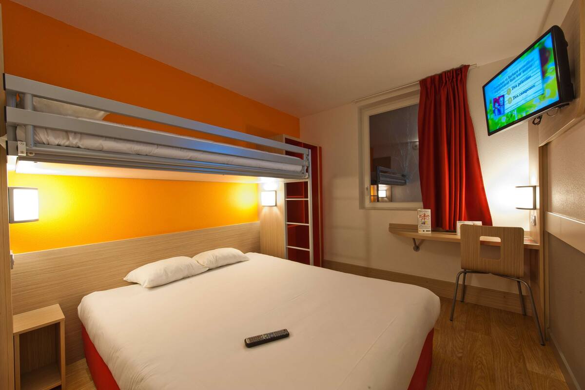 A hotel room with a bunk bed, a desk with a chair, a television mounted on the wall, and a window with red curtains.
