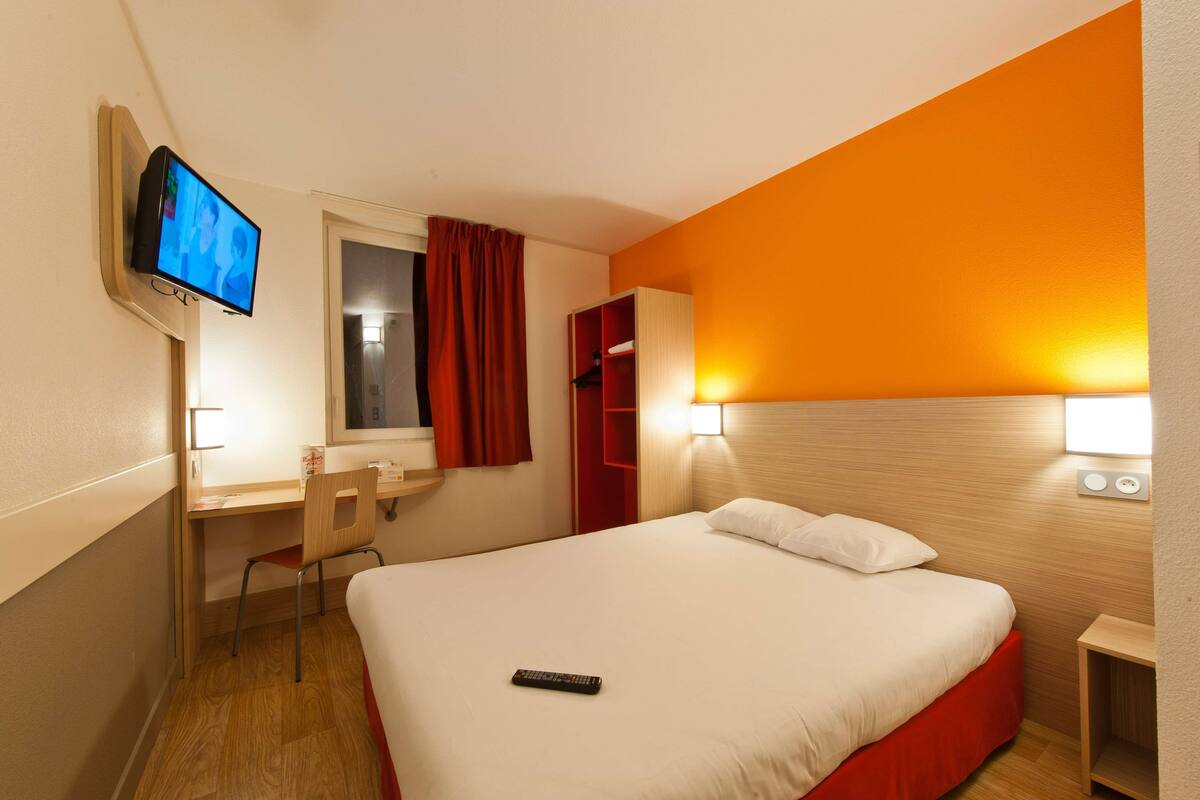A hotel room with a bed, a desk, a chair, a TV mounted on the wall, and a red wardrobe.