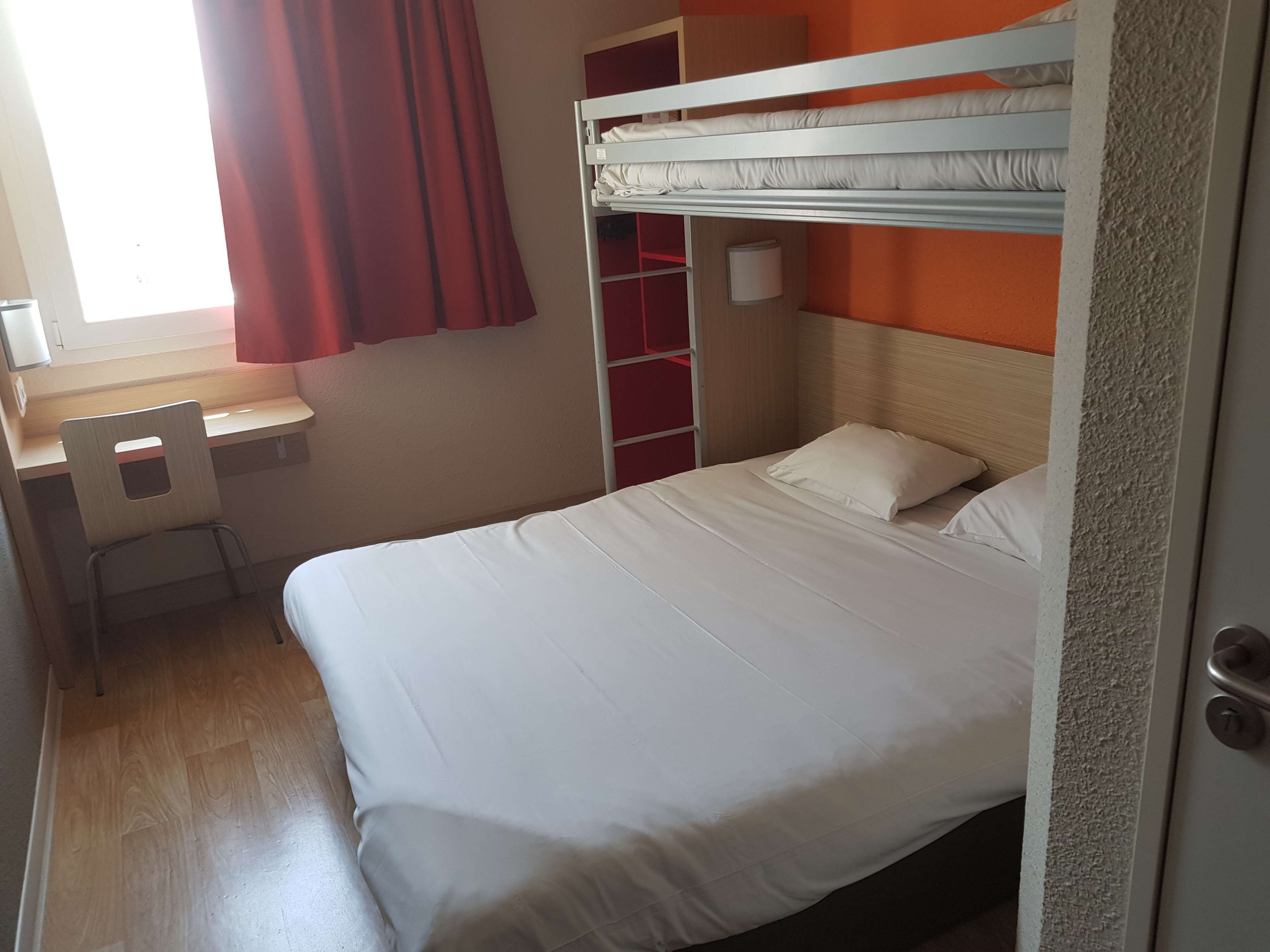 Een slaapkamer met een stapelbed, bureau en stoel, een rood gordijn en een plankunit.
