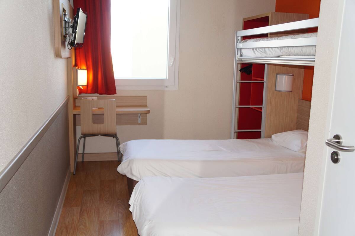 A small, compact hotel room with two single beds, a desk, a chair, and a window with curtains.