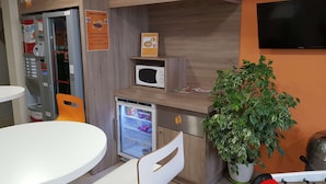 Una kitchenette compacta con microondas, mini refrigerador y pequeño refrigerador. Hay una mesa y una silla, una planta en maceta, y un televisor montado en la pared.