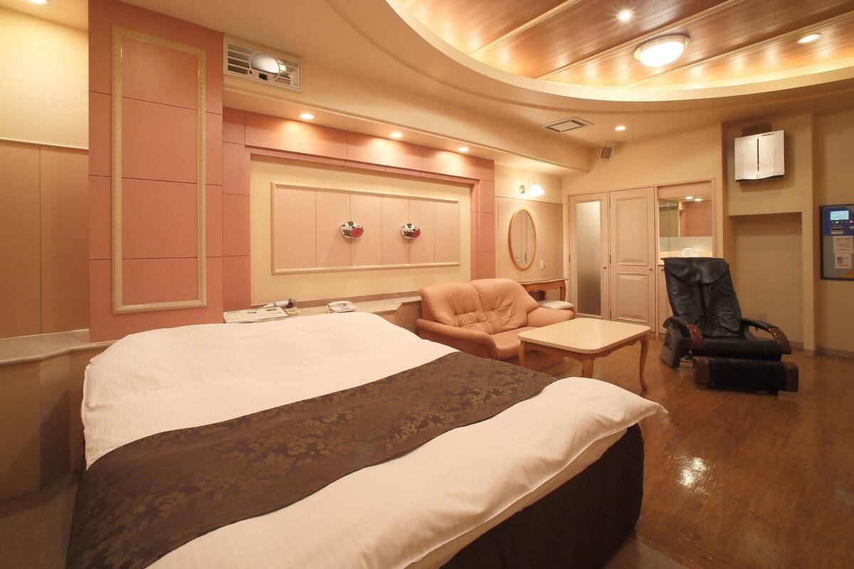 Une chambre d’hôtel comprenant un grand lit, un coin salon avec un fauteuil et une table, une coiffeuse avec un miroir et une armoire.