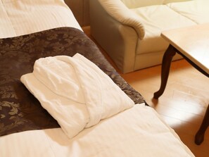 Ein ordentlich bezogenes Bett mit einem weißen Handtuch und einem gemusterten Kopfkissen, ein Holztisch und ein beigefarbener Sessel in einem Zimmer mit Holzboden.