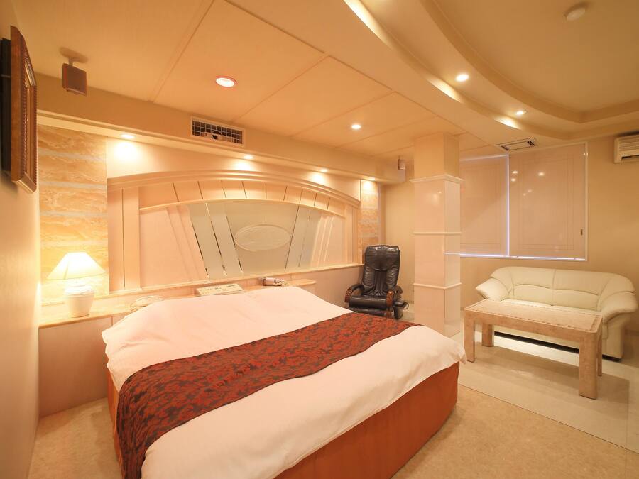 A bedroom with a large bed, a massage chair, a sofa, and a window with blinds.