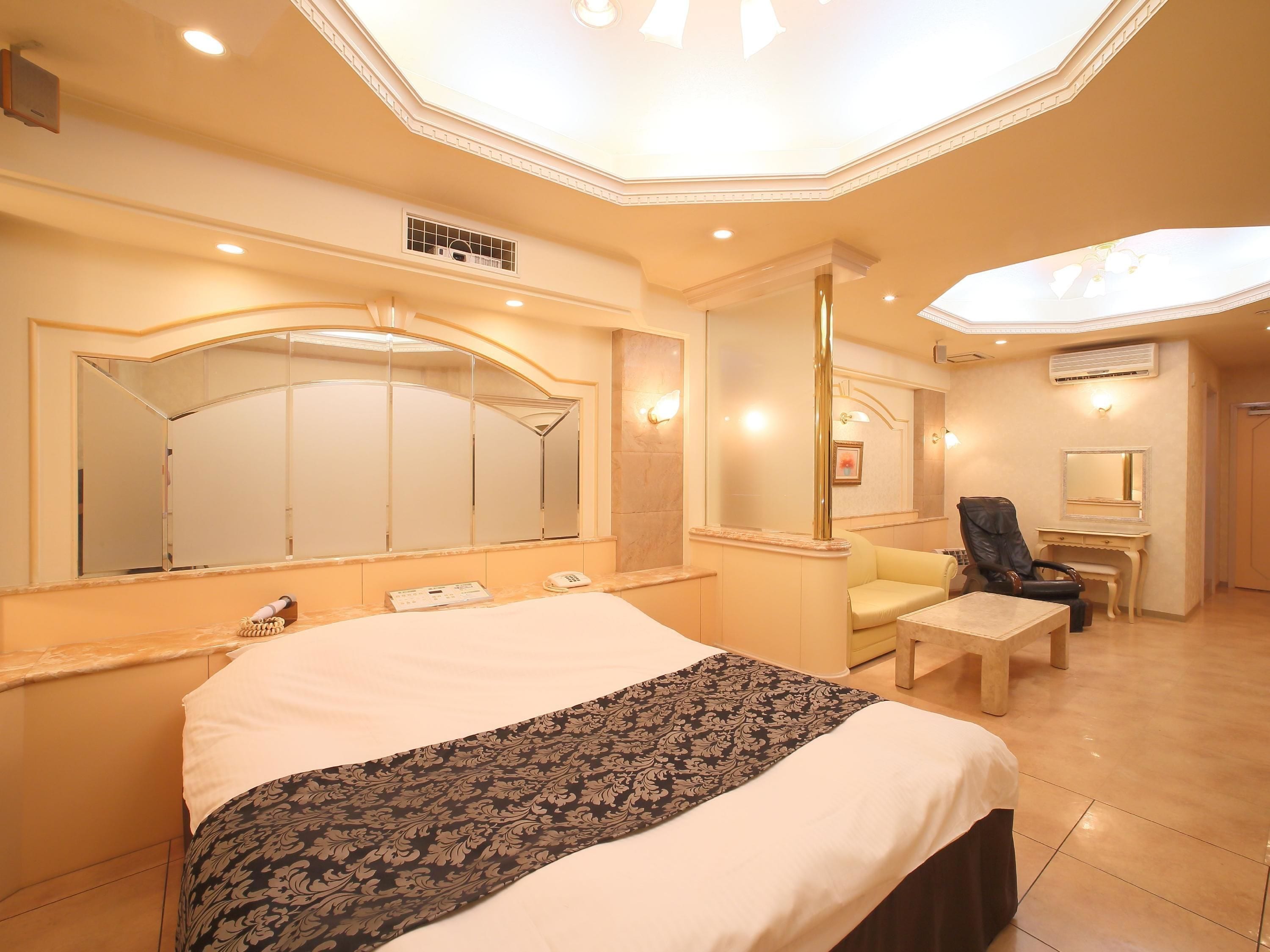 A spacious hotel room with a large bed, a sitting area with a chair and a small table, and a large mirror above the bed.