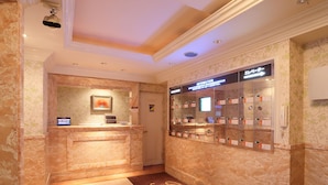 Zona de recepción de hotel con mostrador de mármol, un panel informativo digital, mostrador de recepción y un directorio con números de habitaciones.