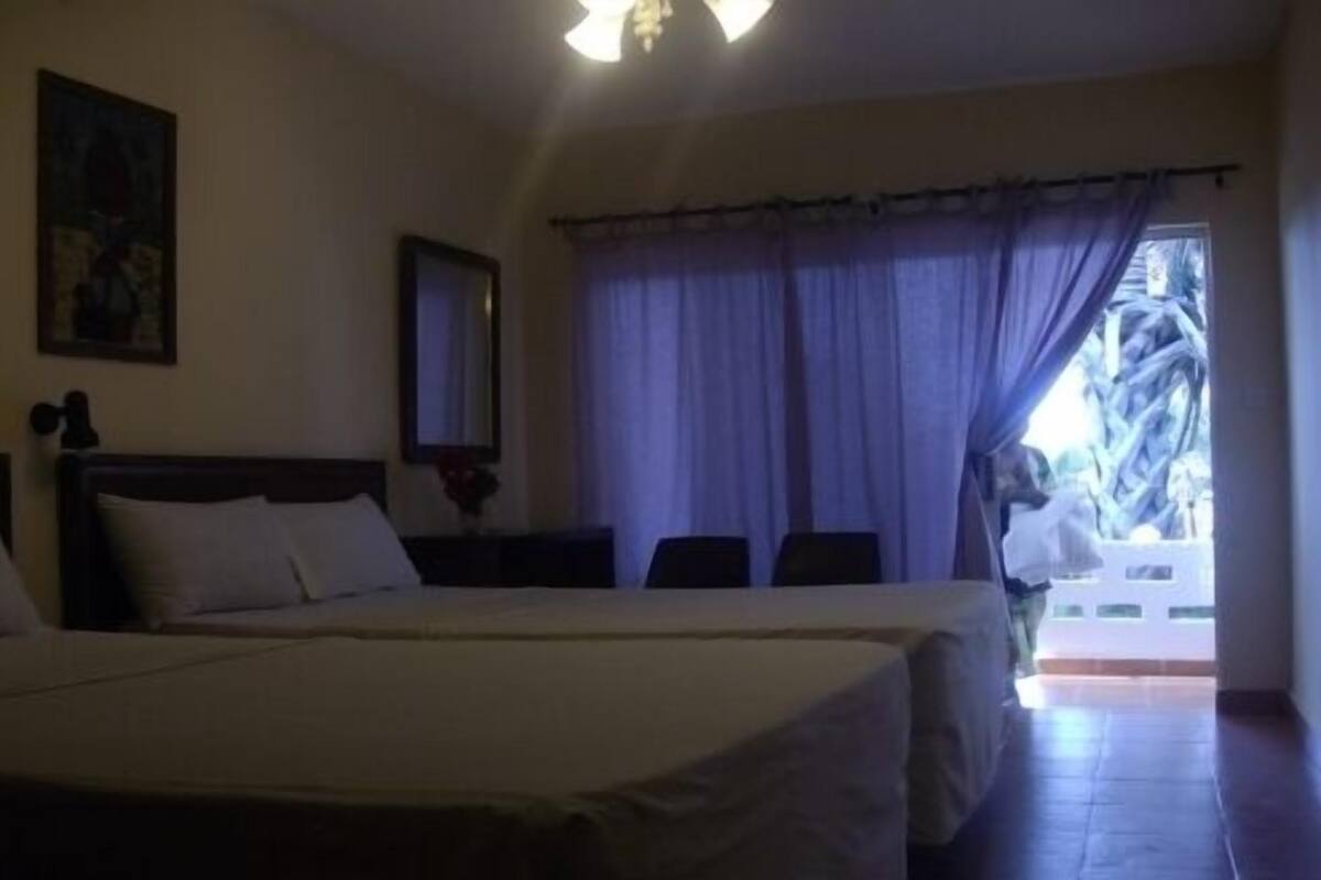 A hotel room with two beds, a large window with curtains, a painting on the wall, and a ceiling light.