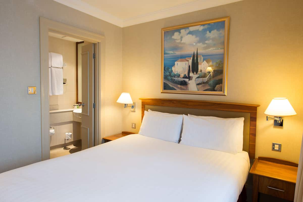 Habitación de hotel con una cama grande, dos lámparas de noche, un cuadro en la pared y un baño visible a través de una puerta abierta.