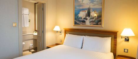 A hotel room with a large bed, two bedside lamps, a painting on the wall, and a bathroom visible through an open door.