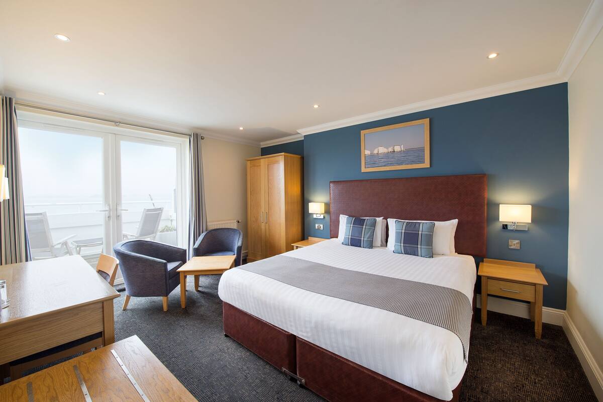 A hotel room with a large bed, a desk, a chair, a wardrobe, and a view of the sea through the windows.