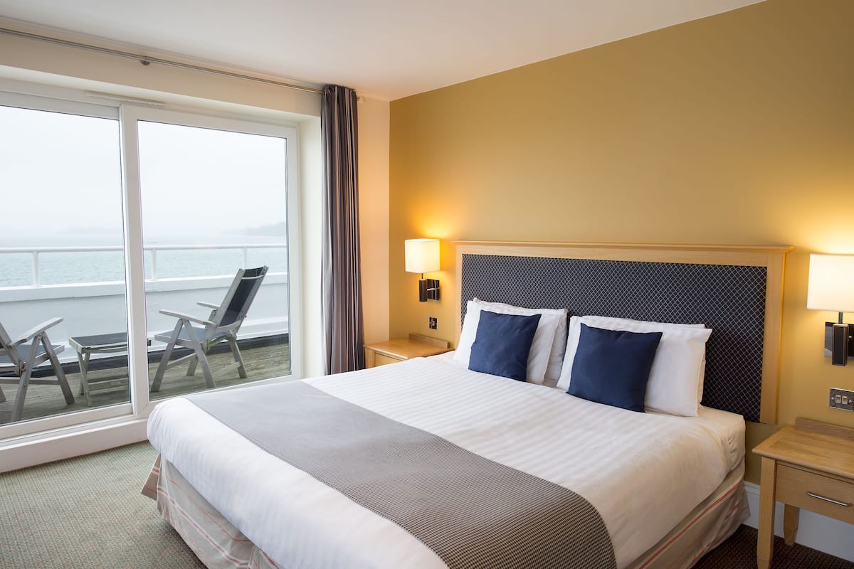 Habitación de hotel con una cama grande, una mesita de noche, una lámpara, una silla y vistas al mar a través de una puerta corrediza de cristal.