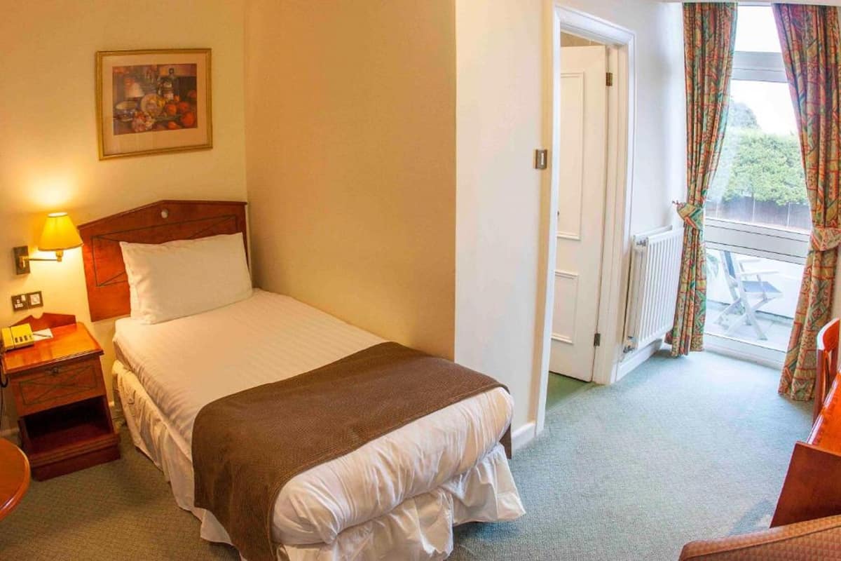 Habitación de hotel con cama, mesita de noche, lámpara y vista al balcón exterior.