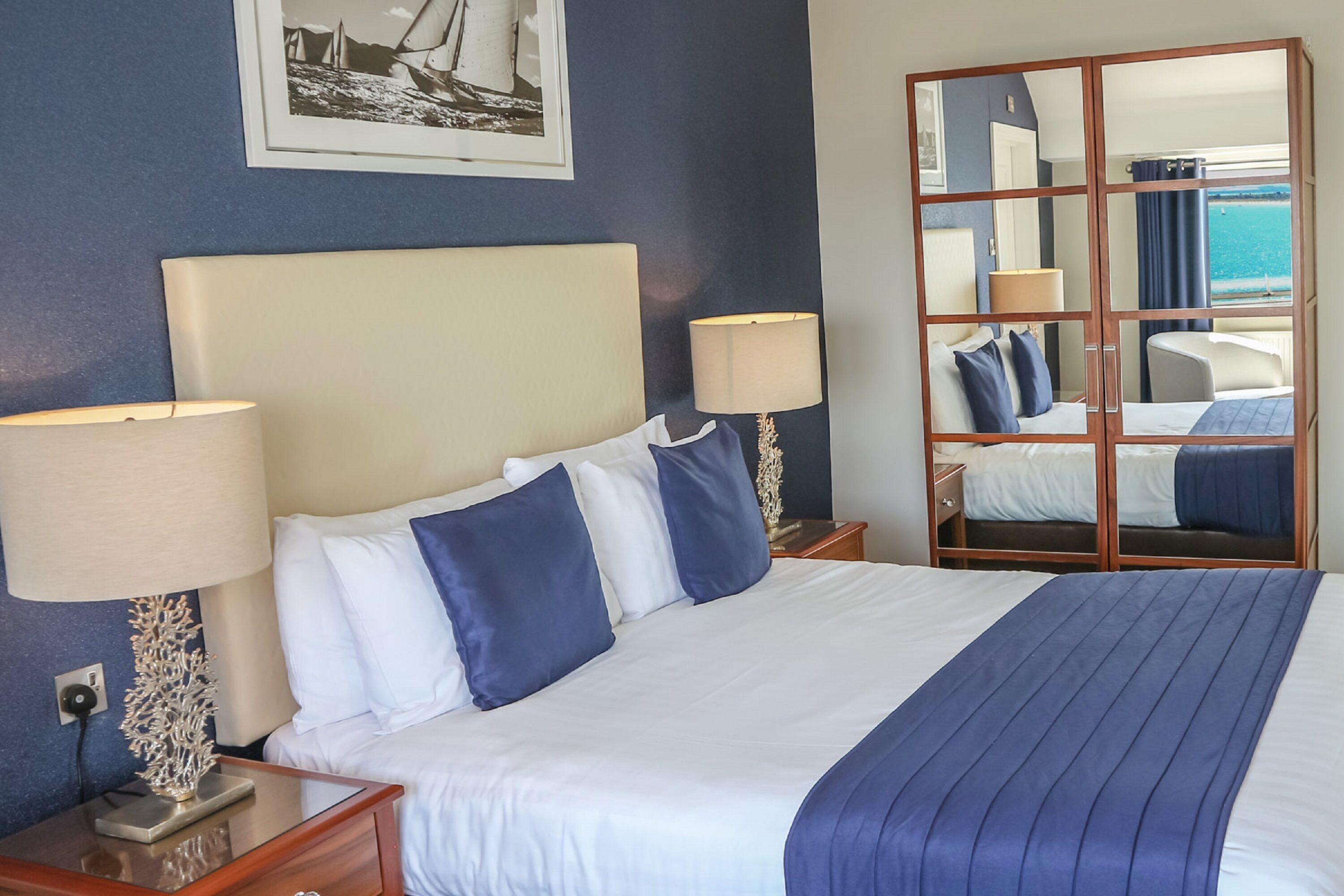 Habitación de hotel con una cama grande, dos lámparas de noche, una mesita de noche, un espejo y vistas al mar.