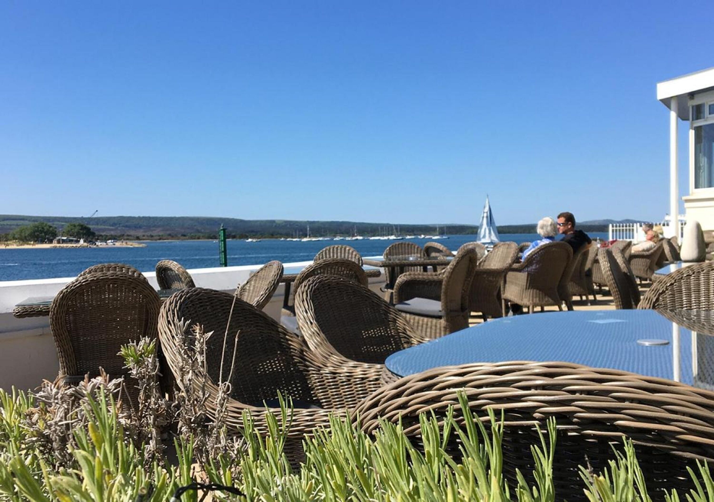 Wicker chairs and tables by a waterfront, with a clear view of the sea and distant hills.