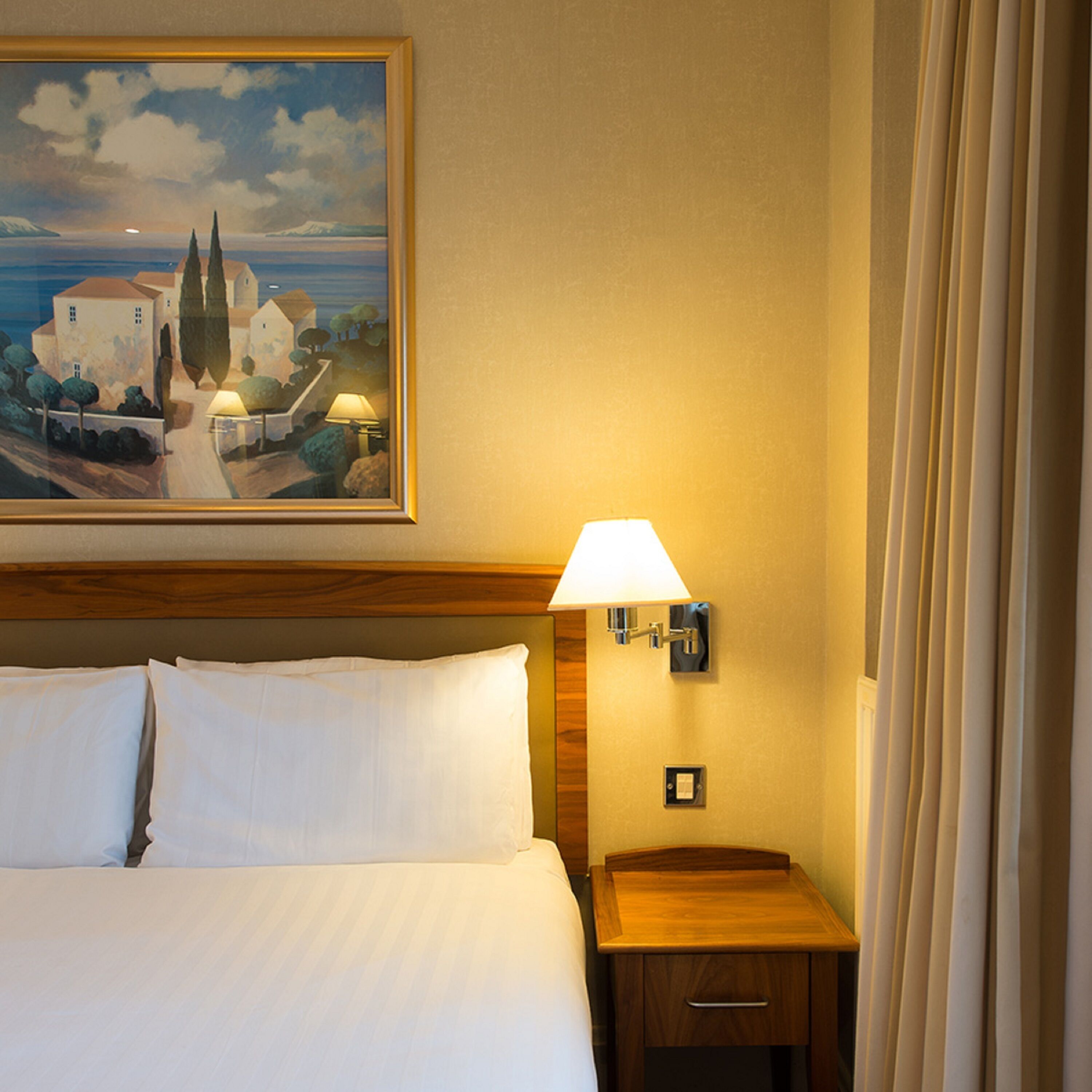 Quarto de hotel com cama, mesa de cabeceira com luminária, pintura de uma paisagem costeira e janela com cortinas.