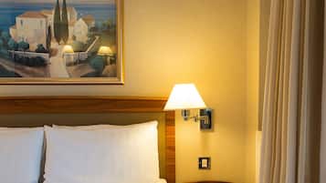 A hotel room with a bed, a nightstand with a lamp, a painting of a coastal scene, and a window with curtains.