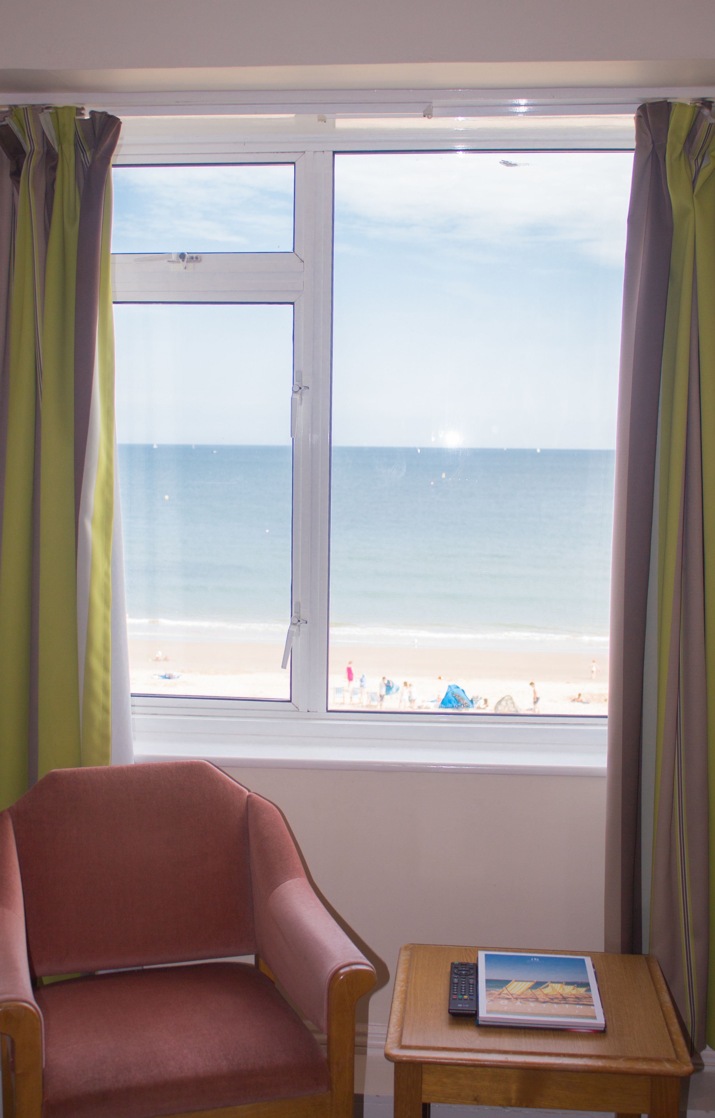 A room with a window view of a beach, a chair, and a small table with a remote control and a book.