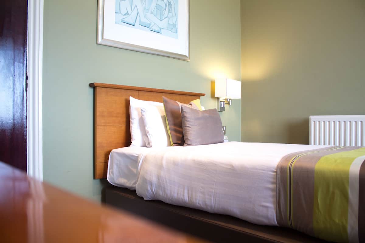 A neatly made bed with a wooden headboard, a framed artwork above it, and a wall-mounted light fixture to the right.