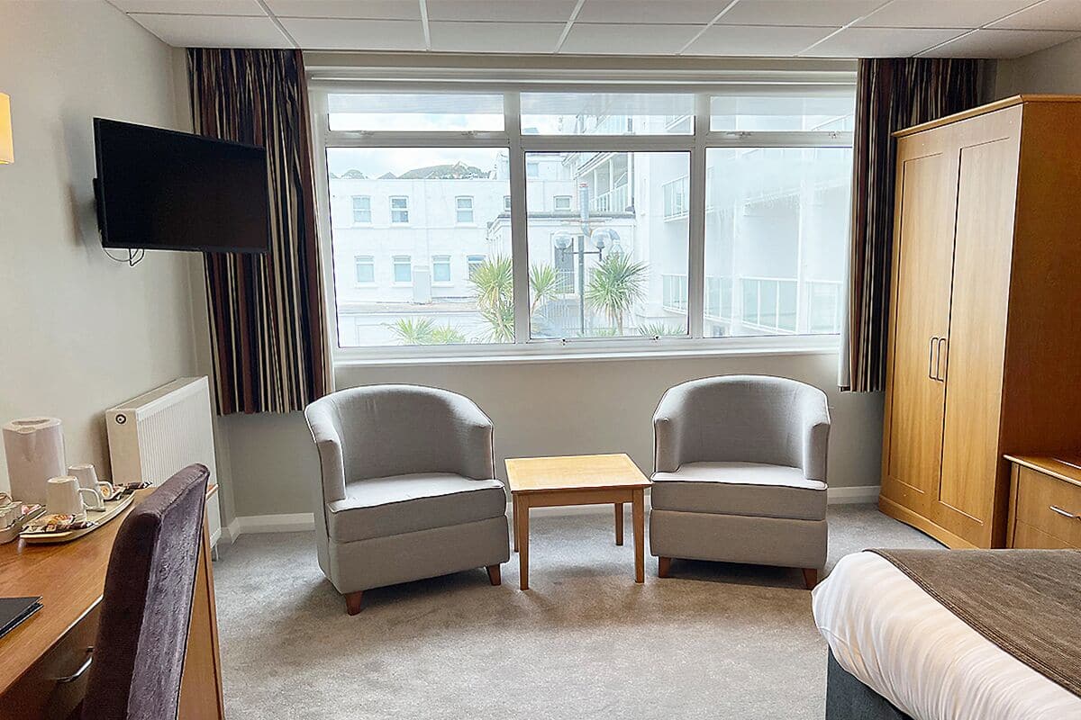 A hotel room with two armchairs, a small table, a bed, a TV mounted on the wall, and a view of the city through large windows.