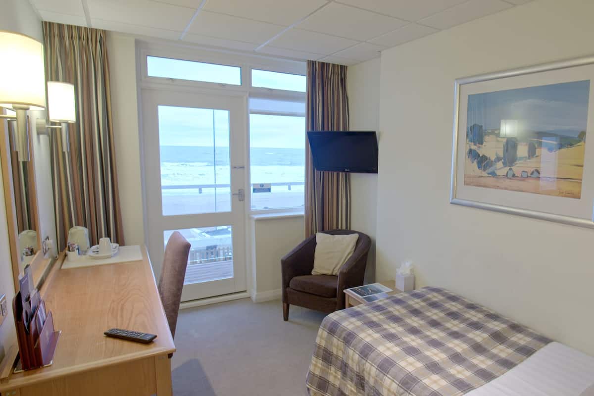 A hotel room with a bed, a desk with a computer, a chair, a TV, and a window with a view of the beach.