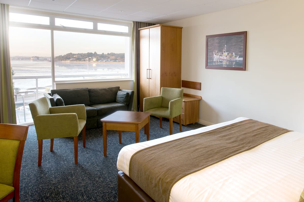 A hotel room with a bed, a sofa, a chair, a small table, and a desk. There is a window with a view of a body of water and buildings.