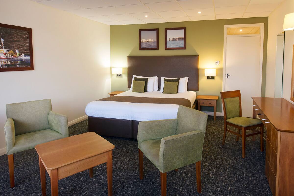 A hotel room with a bed, two armchairs, a wooden table, a desk, and a mirror.