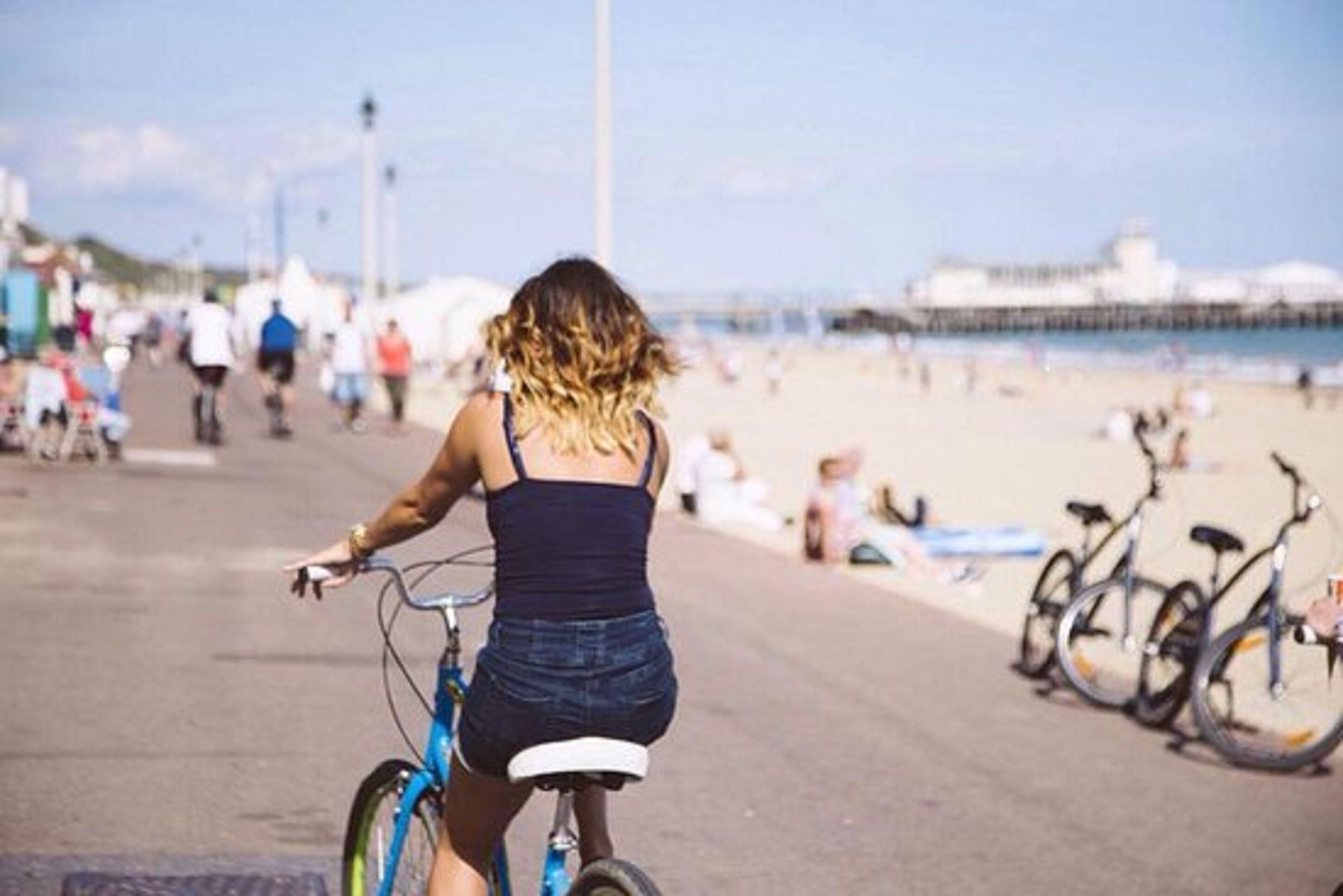 En person cykler på en asfalteret sti tæt på en strand, med cykler parkeret langs siden.