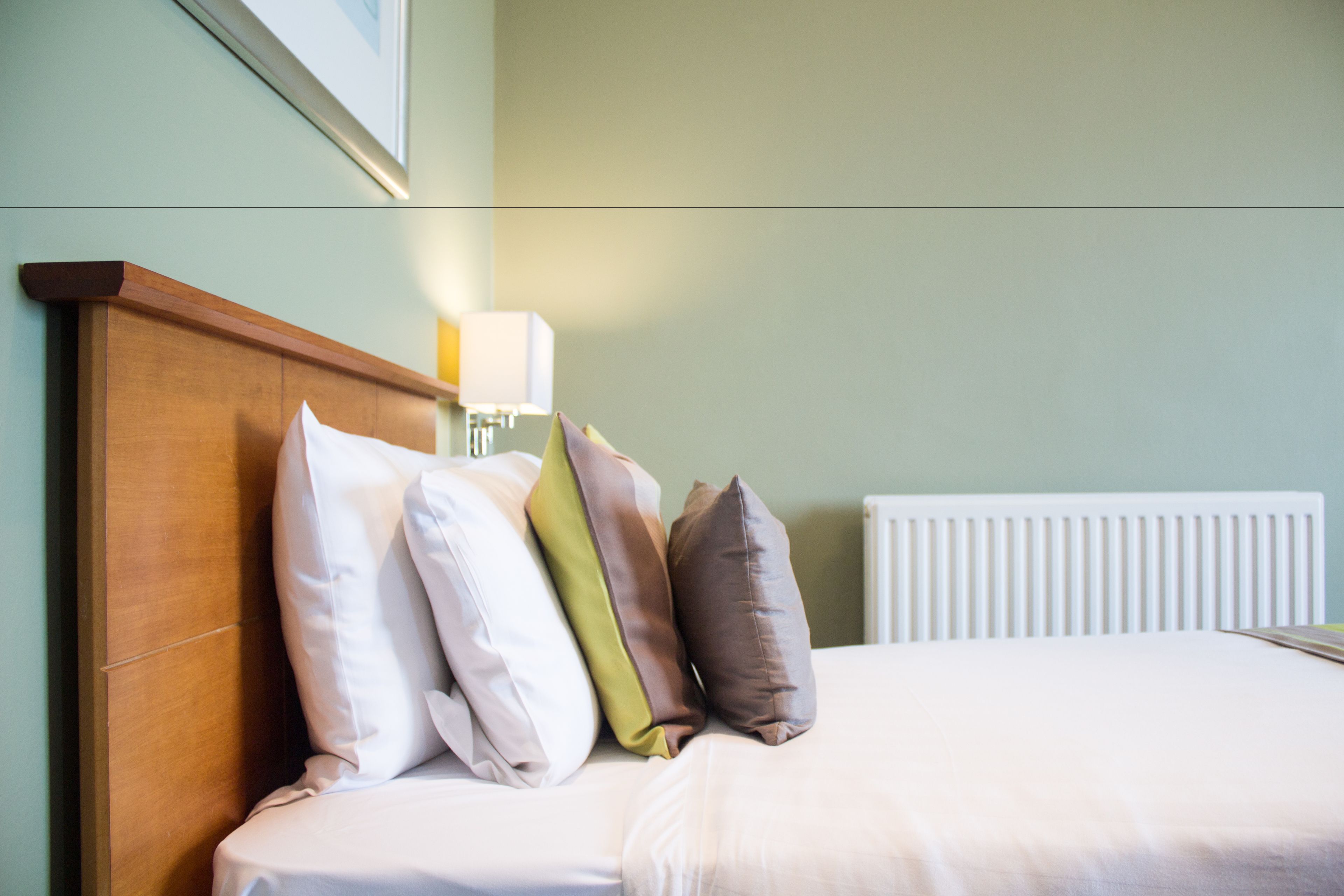 Una cama bien hecha con cabecera de madera, una lámpara de noche y un radiador.