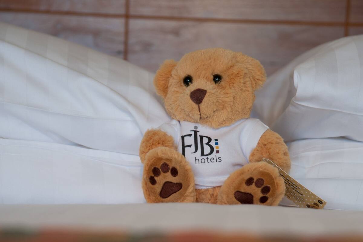 A teddy bear sitting on a bed with a white pillow and a white sheet.