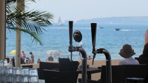 Un restaurante frente al mar con terraza, vista al océano y un velero a lo lejos.