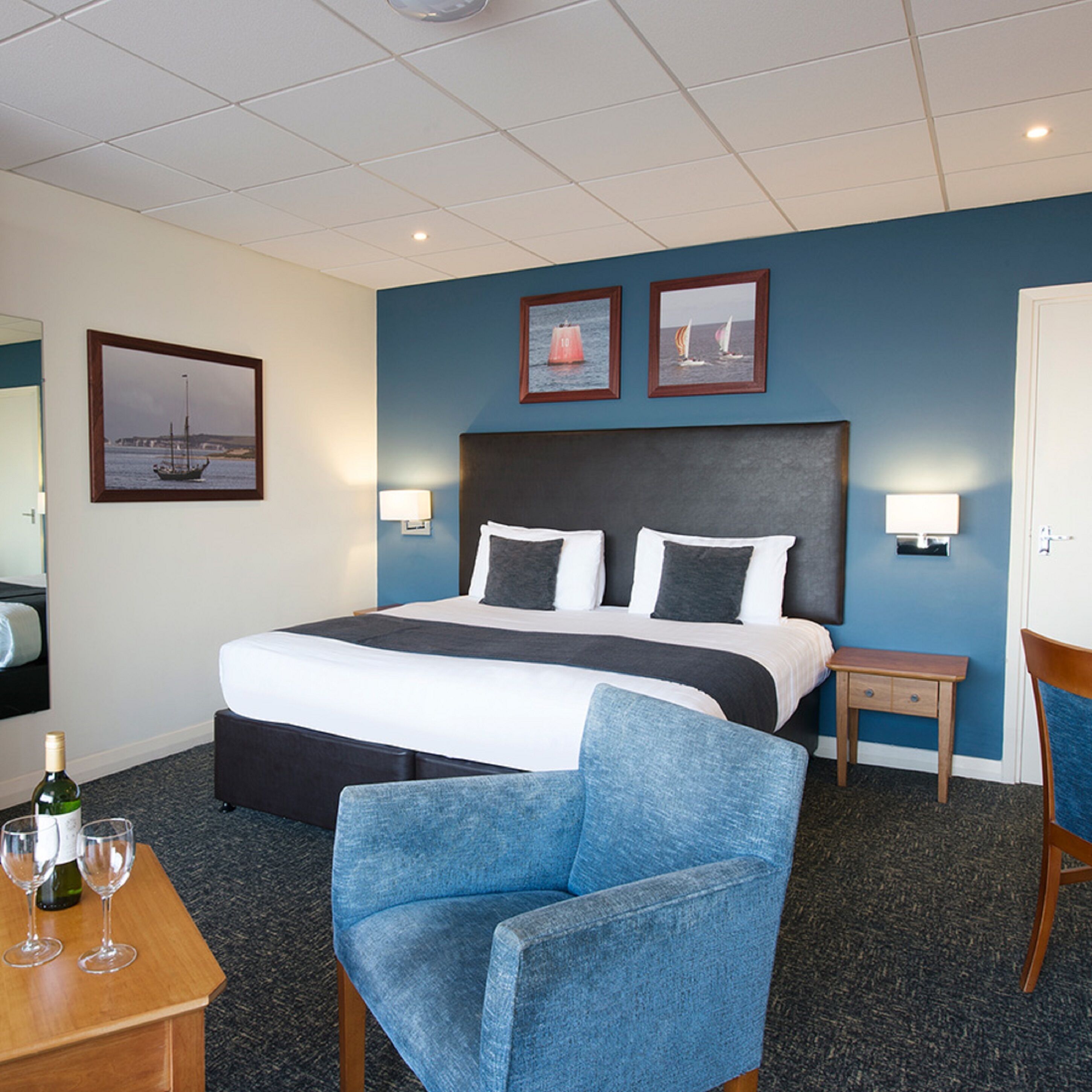 A hotel room with a large bed, a chair, a small table with glasses and a bottle, and two framed pictures on the wall.