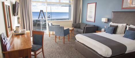 Habitación de hotel con una cama grande, un escritorio con silla, una mesita y vistas al océano a través de amplias ventanas.
