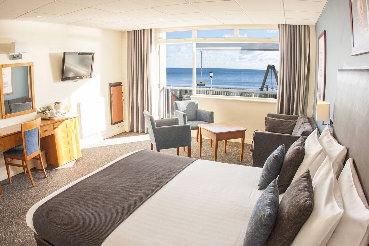 A hotel room with a large bed, a desk with a chair, a small table, and a sofa. There is a TV mounted on the wall and a window with a view of the sea.