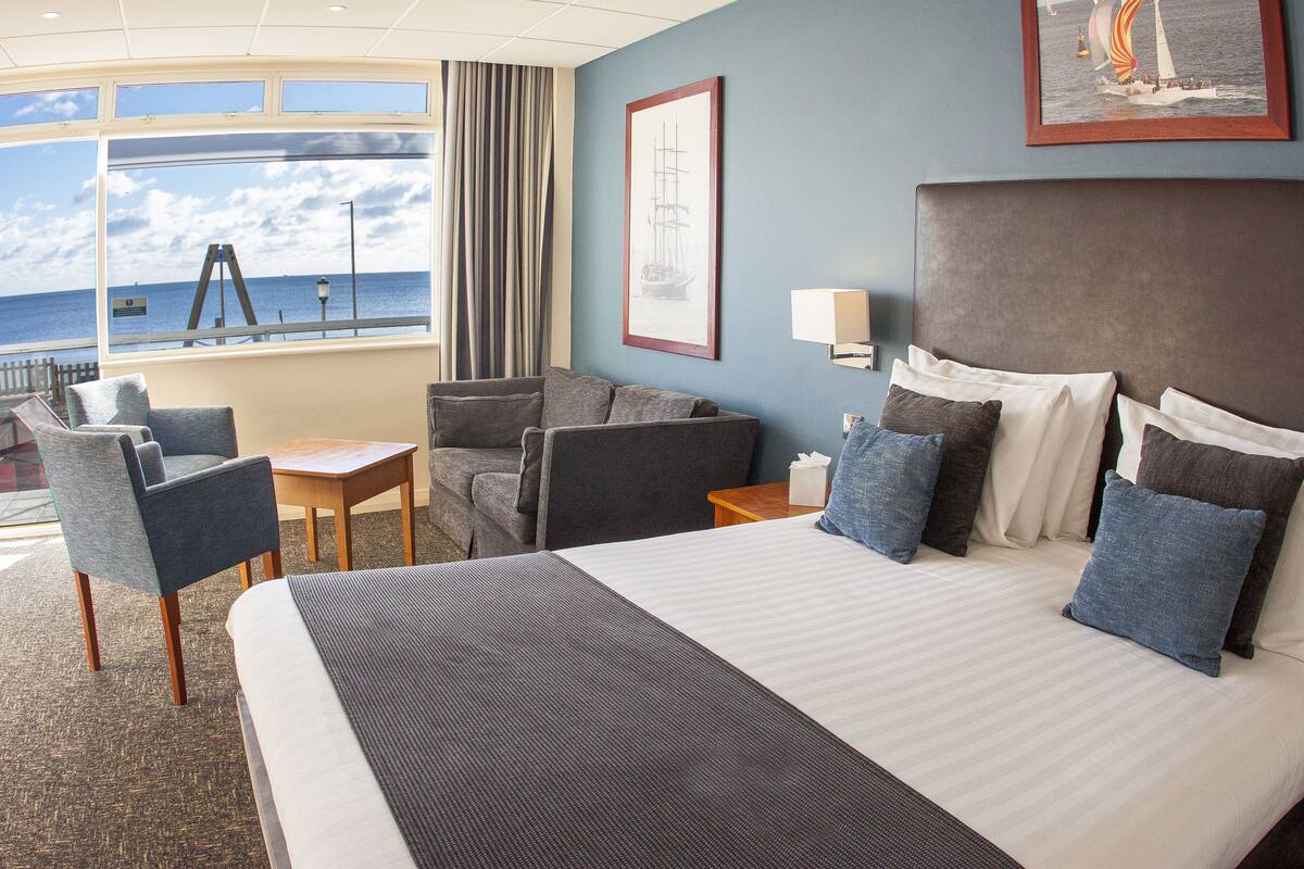 A hotel room with a large bed, two armchairs, a small table, and a view of the sea through a large window.
