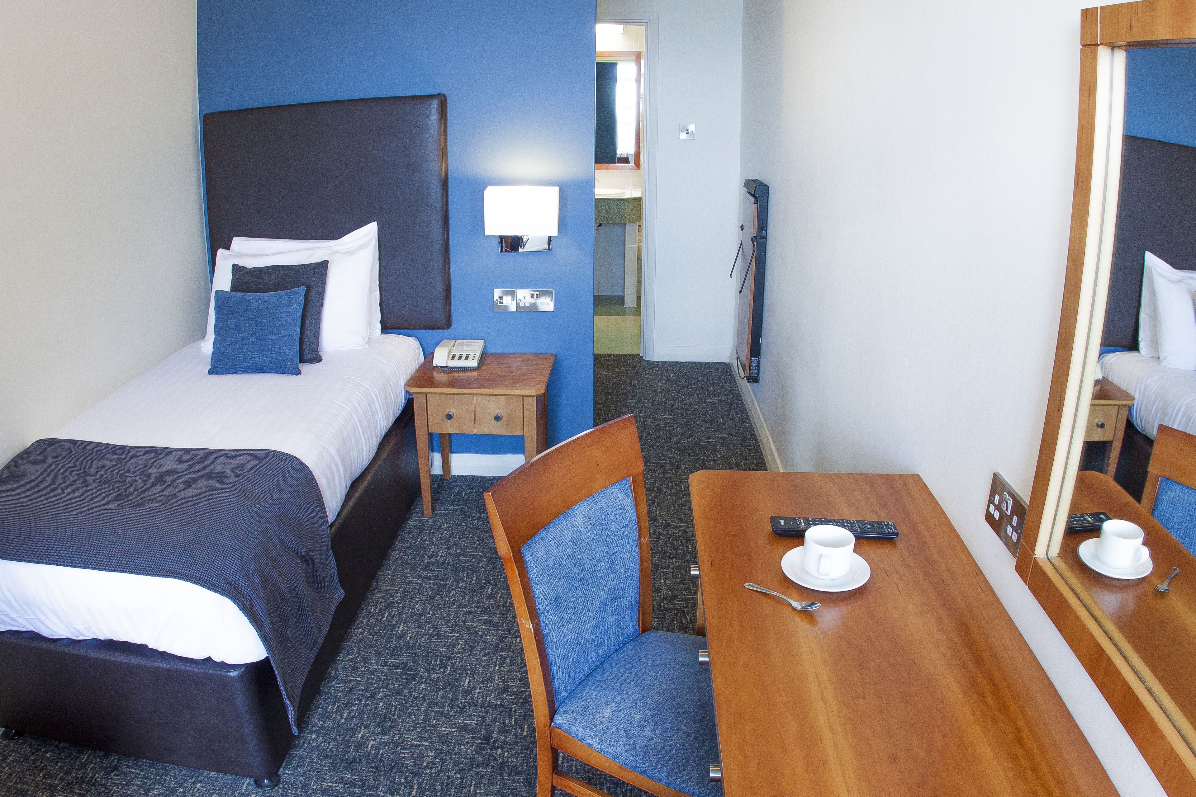 Une chambre d’hôtel avec deux lits, un bureau en bois, une chaise, un miroir et une petite table sur laquelle se trouvent une tasse et une télécommande.