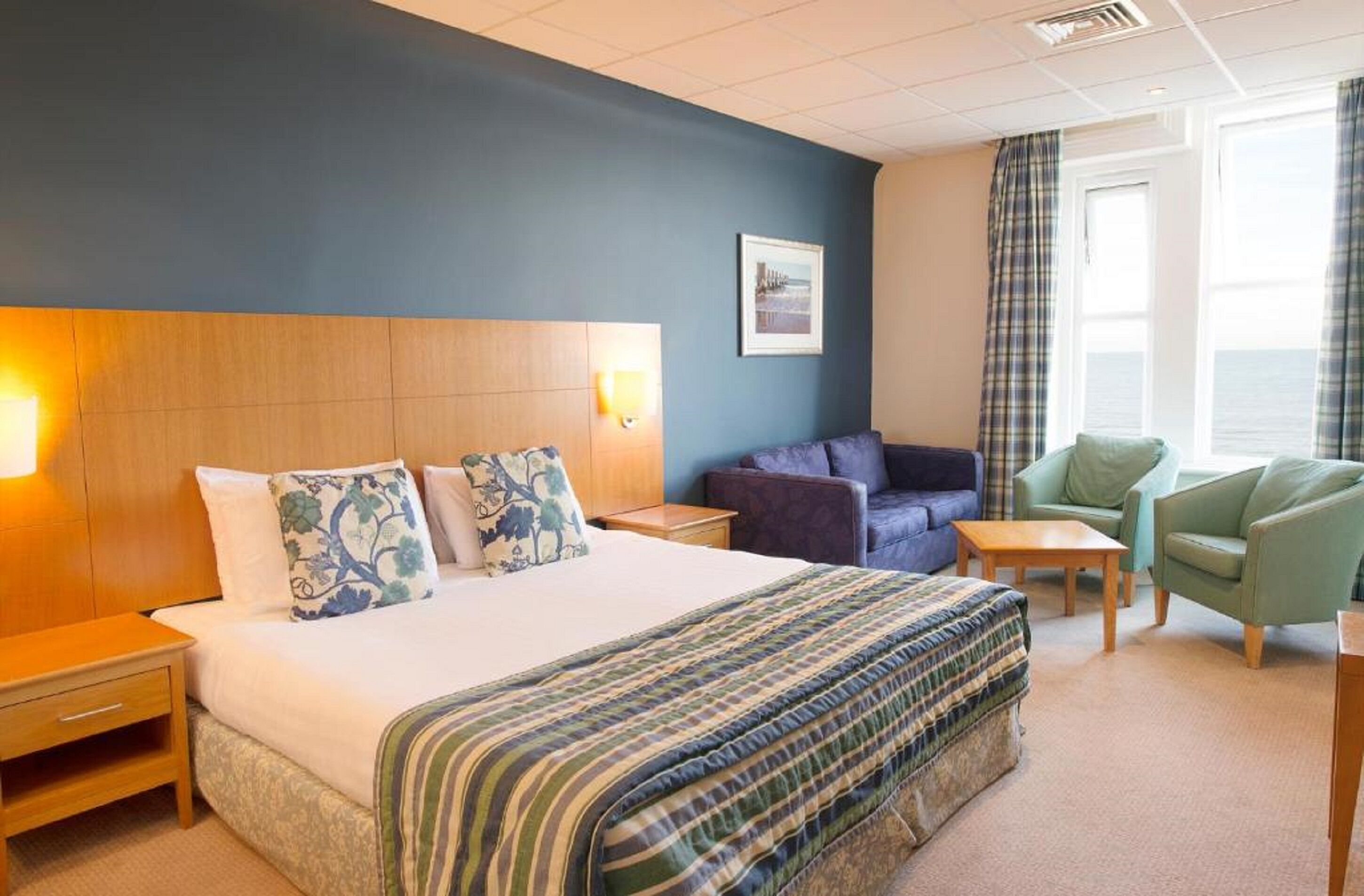 A hotel room with a large bed, a sofa, two armchairs, a small table, and a window with curtains.
