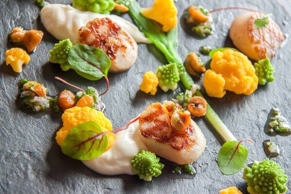 Primer plano de un plato gourmet con vieiras selladas, verduras y salsa cremosa.