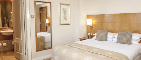 A hotel room with a large bed, a wooden headboard, a bedside table with a lamp, a mirror, and a bathroom with a sink and toilet.