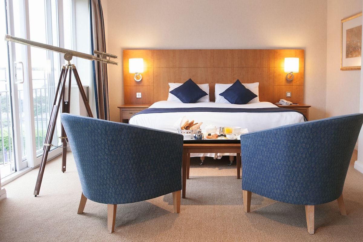 Ein Hotelzimmer mit Bett, Nachttischlampen, einem Schreibtisch, einem Sessel und einem Fenster.