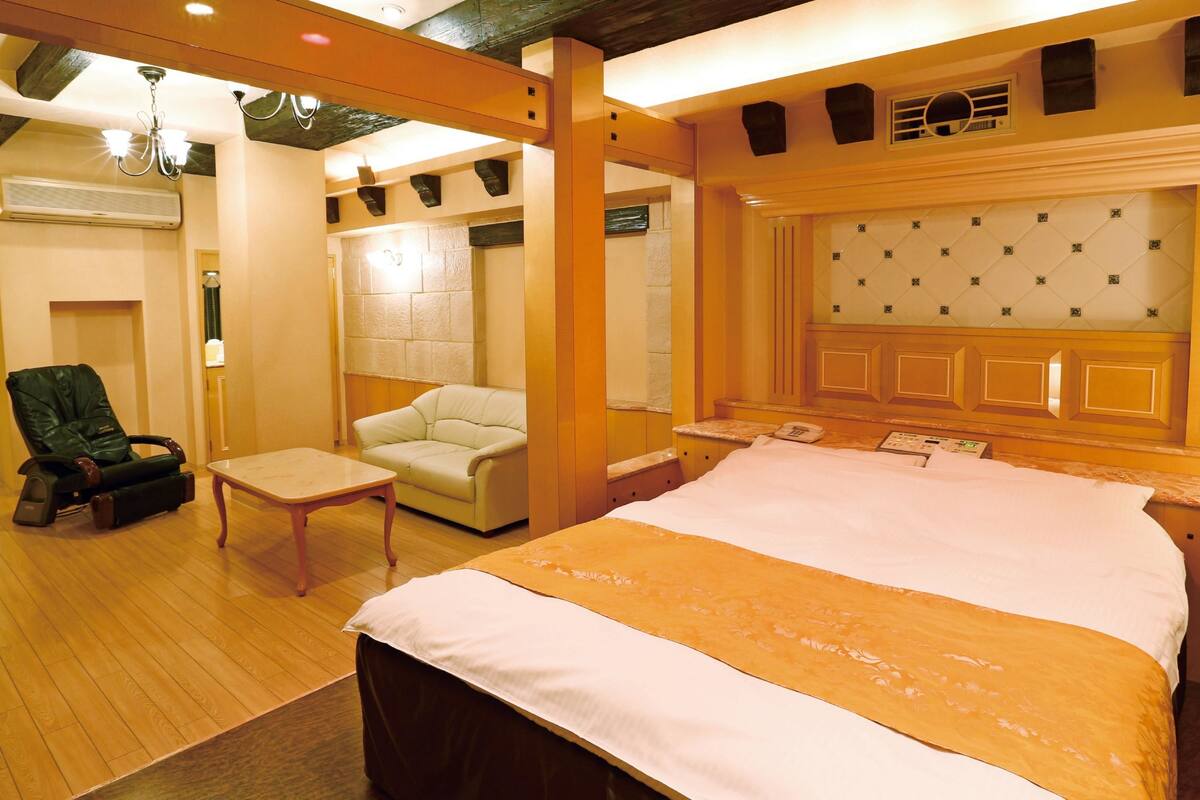 A hotel room with a large bed, a massage chair, a sofa, and a small table.