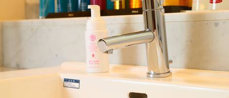 A bathroom sink with a modern faucet, a bottle of facial soap, and various toiletries in the background.