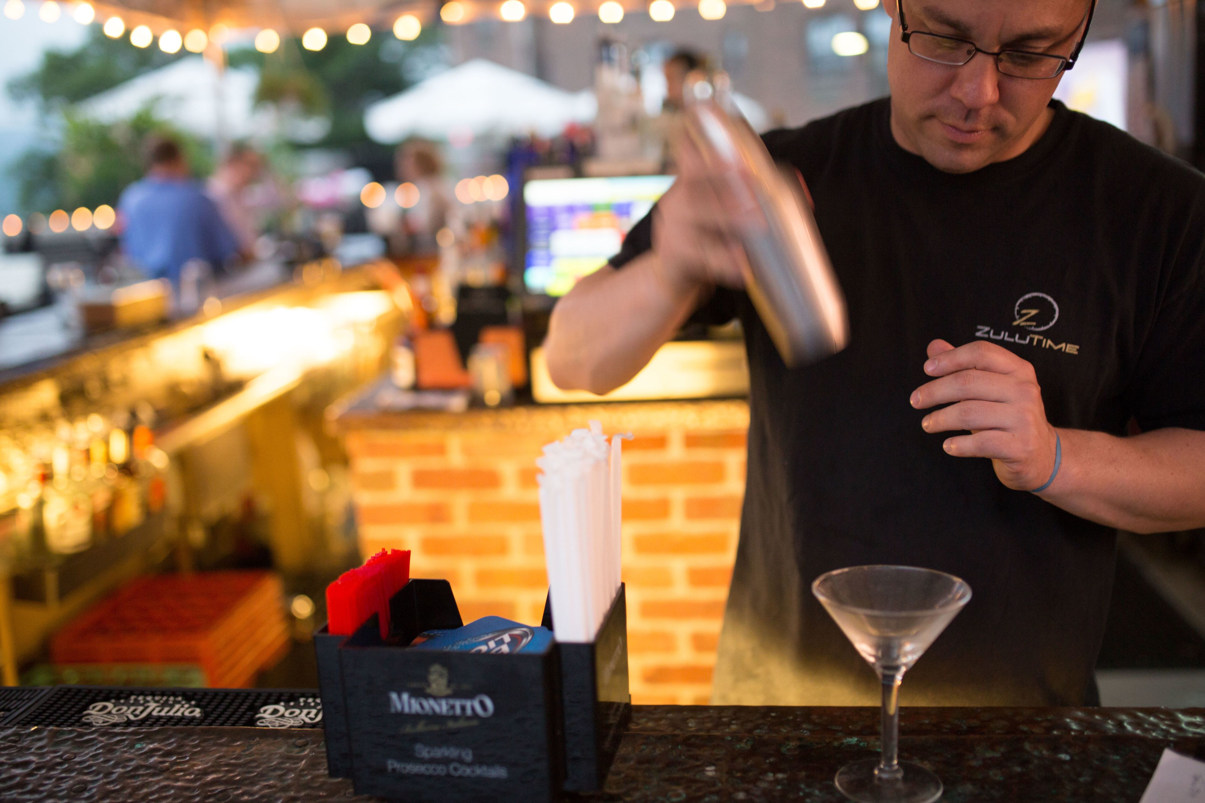 Un cantinero prepara una bebida en la barra, con un fondo de pared de ladrillo y una botella de Martini.