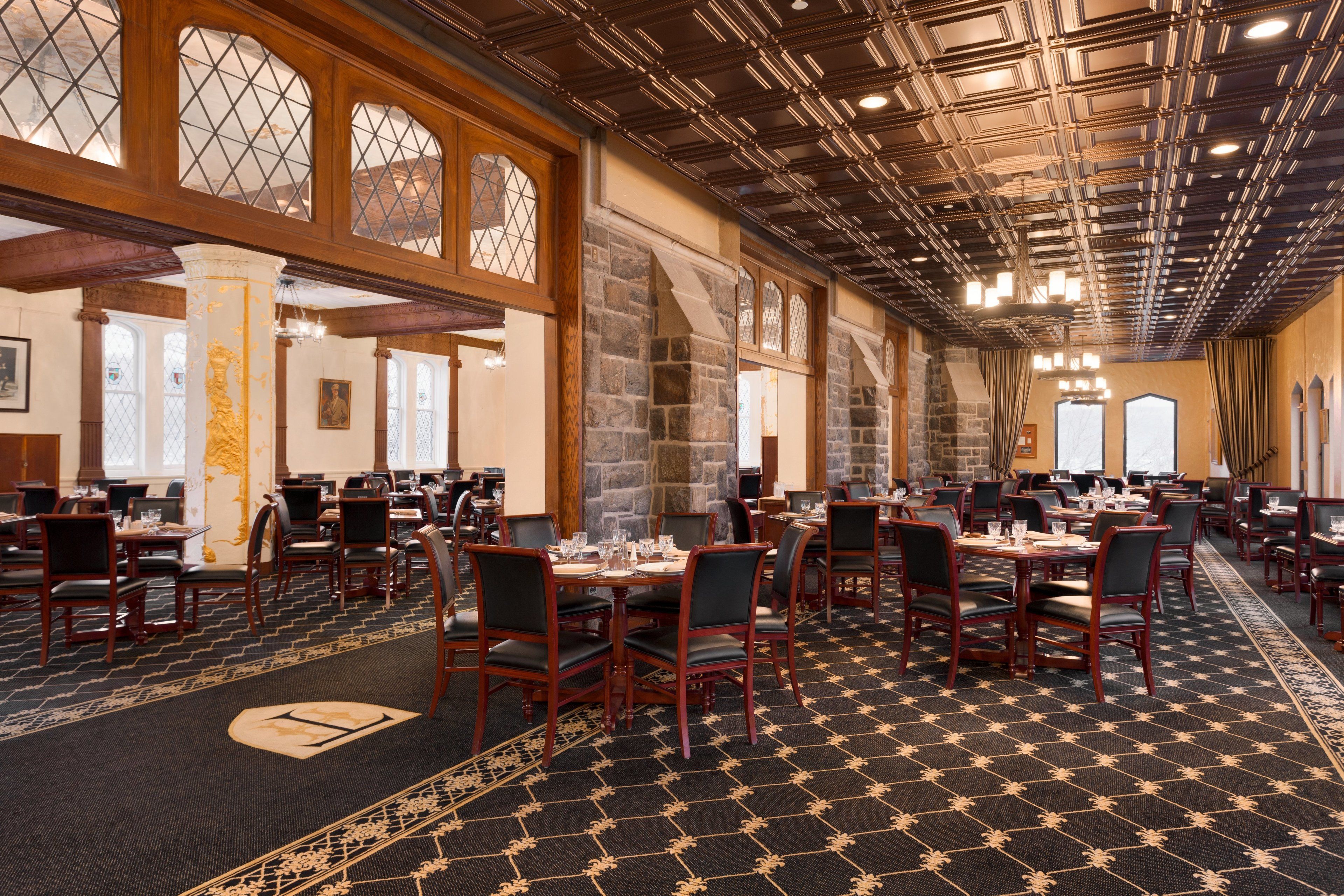 A spacious hotel dining area with multiple tables and chairs, large windows, and a patterned carpet.